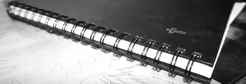 an black and white image of a dark spiral bound journal with a silver tinted sticker to the right side of its cover in the shape of a Christian cross